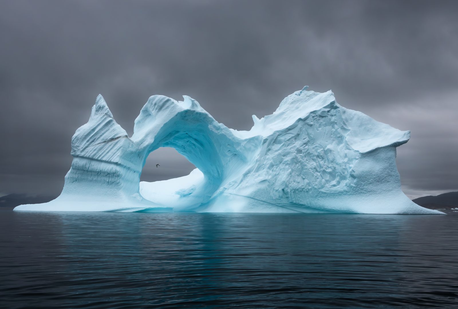 An arched iceberg of pale blue ice standing in still dark water under a heavy grey sky, Greenland.